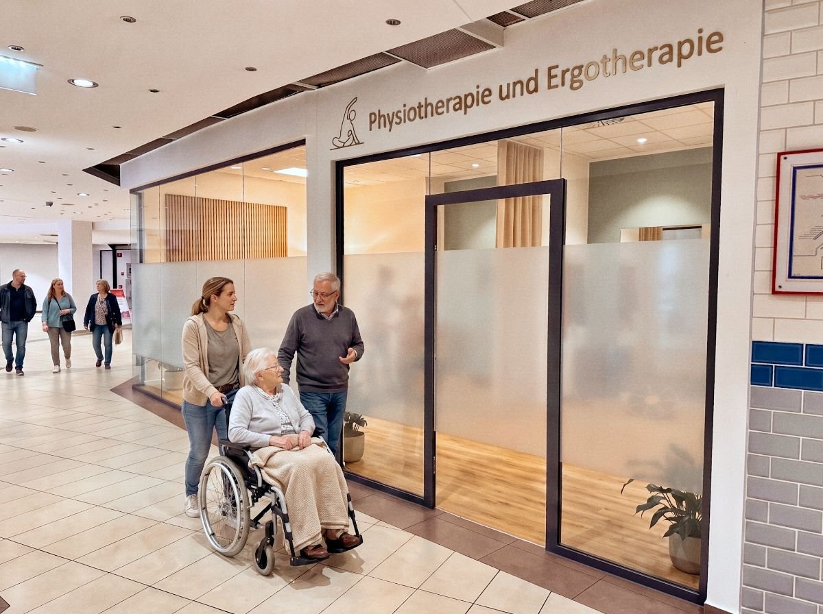 Eine Frau schiebt eine ältere Dame im Rollstuhl vor der Physio- und Ergotherapie im Porcia MedCenter, ein Mann geht daneben